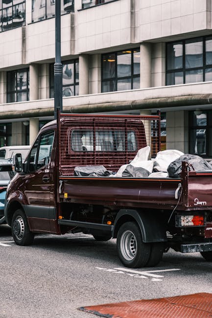A small burgundy flatbed truck parked on an urban street in front of a modern office building with large glass windows and concrete facade. The truck's cargo bed is loaded with various types of waste, including black and white garbage bags, cardboard boxes, and loose debris, indicating a recent rubbish removal. The vehicle features a protective metal grille behind the cab to secure the load and has visible side mirrors and standard steel wheels. The surrounding environment includes a streetlamp and other parked vehicles, with a clear, overcast sky reflected in the building's windows. This scene exemplifies independent waste collection services, such as those provided by Rubbish Clearance Hammersmith, aimed at commercial waste removal for shops and offices, emphasizing professional non-local authority rubbish disposal methods that support alternative waste handling solutions in urban settings.