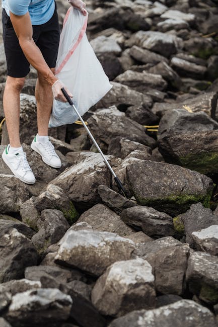 A person wearing a light blue t-shirt, navy shorts, and white sneakers is crouched down on a rocky shoreline, using a grabber tool to collect debris or rubbish among large, irregularly shaped stones with rough textures and dark grey and brown tones. The individual is holding a white trash bag, partially visible, for rubbish collection in an outdoor natural environment, with rocks extending into the background. The scene suggests an activity related to cleaning or waste removal in a rugged, outdoor setting, and the presence of the grabber tool and trash bag indicates independent rubbish collection, similar to private waste disposal activities managed by companies such as Rubbish Clearance Hammersmith, which specialises in local rubbish removal services.
