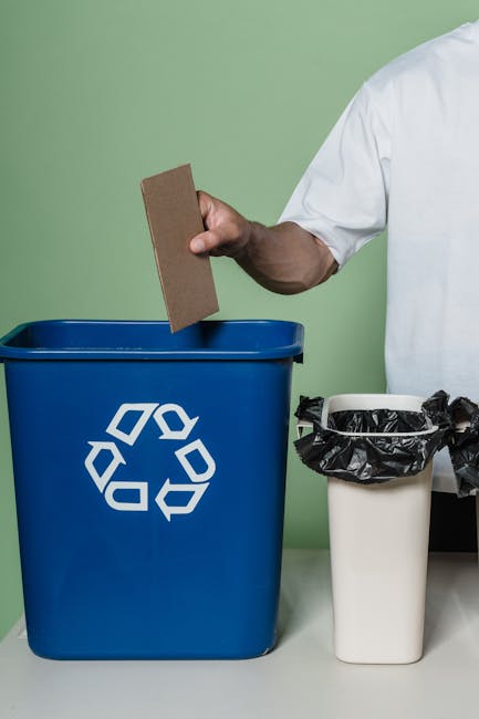 A person in a white shirt is holding a rectangular piece of brown cardboard or recycled paper above a blue recycling bin that features a white recycling symbol on its front. The blue bin is positioned on a light-colored table or countertop, with its opening facing upward. To the right of the blue bin is a smaller white container lined with a black garbage bag, which is loosely gathered over the edges. The background features a plain, light green wall, and the overall scene appears to depict an indoor setting related to waste separation or rubbish collection, emphasizing the process of sorting waste materials for proper disposal or recycling, as part of a rubbish removal service or on-site clearance activity by Rubbish Clearance Hammersmith.