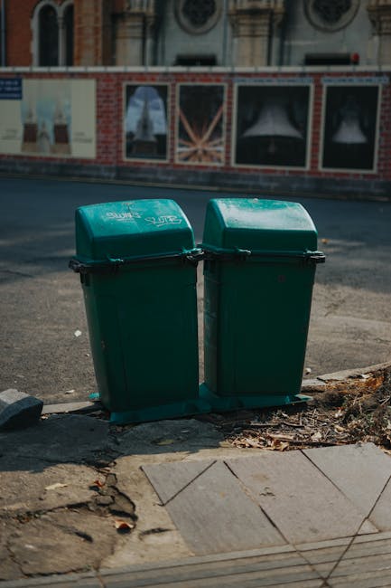 Two large grey wheeled rubbish bins positioned on a paved outdoor area beneath a metal fire escape staircase. The bin on the left is marked with a label indicating it is for 'RECYCLABLE WASTE,' while the one on the right has a label for 'RESIDUAL WASTE.' The bins have textured, plastic surfaces with visible lids and wheels, and are placed against a light grey wall with horizontal siding. Shadows of nearby trees are cast onto the wall, creating a pattern. In the background, part of a brick building, a streetlamp, and some distant rooftops are visible under a clear blue sky, indicating a typical urban environment. This scene, associated with rubbish collection and waste management, aligns with services like those provided by Rubbish Clearance Hammersmith, supporting alternative waste handling methods such as on-site disposal and private clearance.
