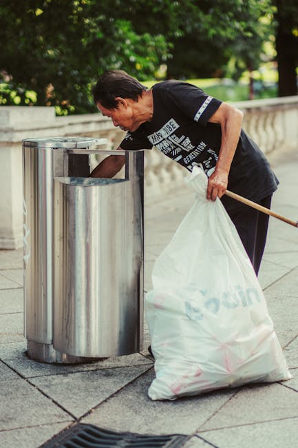 A man with dark hair, wearing a black t-shirt with white text and dark trousers, is seen bending over a stainless steel rubbish bin with three separate compartments, set on a paved sidewalk. He is holding a large white rubbish bag with one hand and using the other hand to open the top of the bin, which is part of an outdoor waste disposal area. The scene is situated in a green, leafy environment, with trees and a park-like setting in the background, and a low stone wall bordering the walkway. The man appears to be engaged in rubbish collection or waste clearance, possibly related to private waste handling or outdoor rubbish removal services. The stainless steel bins have a smooth, reflective surface, contrasting with the rough texture of the paving stones. Bright natural light illuminates the scene, highlighting the clean lines of the waste containers and the greenery surrounding the area, reflecting the typical setting for independent rubbish clearance in an urban or suburban environment, aligned with waste management practices such as those offered by Rubbish Clearance Hammersmith.