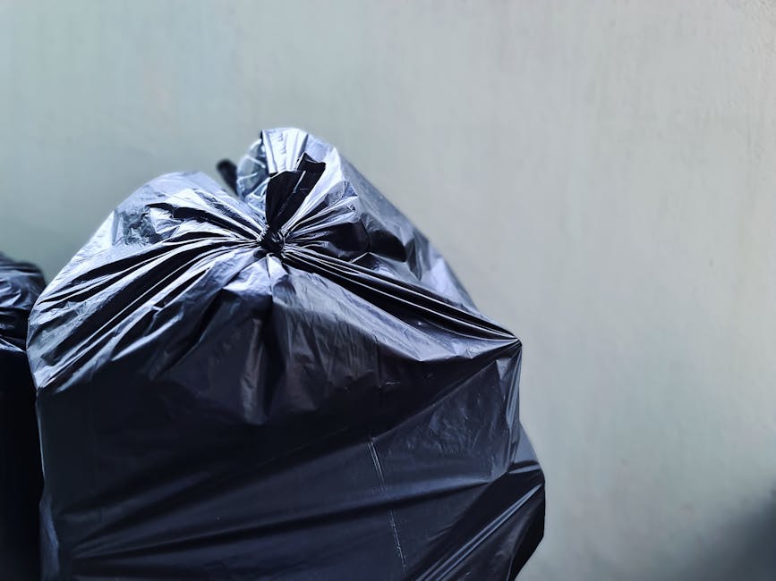 Close-up image of tightly tied black plastic rubbish bags filled with waste, positioned on a smooth, light-colored surface in front of a plain, neutral-toned background. The bags have a glossy texture, with visible creases and folds, indicating they are filled with household or general waste. The top of the bag in the foreground is gathered and securely knotted, showing slight deformation from the contents inside. Additional rubbish bags are partially visible behind it, slightly out of focus. The scene appears to be set outdoors or in a utility area, with natural lighting illuminating the bags uniformly, emphasizing their reflective surface. This visual context aligns with the services offered by Rubbish Clearance Hammersmith, illustrating the typical appearance of waste ready for collection or disposal as part of private rubbish clearance or alternative waste handling solutions.
