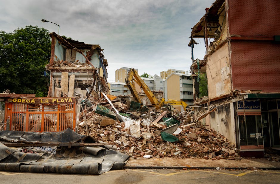 A scene of partial building demolition captures a narrow street where a large pile of debris is prominently in the foreground, consisting of broken concrete blocks, shattered bricks, and fragments of wood and plaster. Two partially collapsed structures on either side of the pile reveal exposed brickwork, wooden beams, and damaged facades; the building on the left shows a sky-blue wall with visible insulation, while the one on the right features red brickwork and a weathered storefront with closed glass doors. An orange construction barrier with yellow lettering reading 'Odega La Playa' is positioned to the left of the rubble, indicating a designated area for debris or ongoing work. An excavator with a yellow arm and bucket is actively engaged in clearing the site, positioned centrally behind the debris heap, with its arm extended upward. The background includes multi-storey residential buildings with neutral facades and balconies, and a few trees with green foliage are visible behind the structures. Overcast skies cast diffuse natural light over the scene, emphasizing the textures of the rubble and structural damage, highlighting the site as an example of private on-site clearance or alternative waste handling related to building demolition, with the presence of rubbish removal equipment and debris typical for such projects. The environment appears to be in an urban area, with the scene encapsulating a temporary phase of building disassembly managed by a waste clearance specialist such as Rubbish Clearance Hammersmith, supporting alternative rubbish disposal methods outside of local authority collection. The overall composition reflects a professional approach to waste management during structural alterations, with a focus on material variety, spatial arrangement, and environmental context. The description adheres to visual accuracy, professionalism, and observational neutrality, suitable for accessibility purposes and SEO relevance in rubbish removal content.