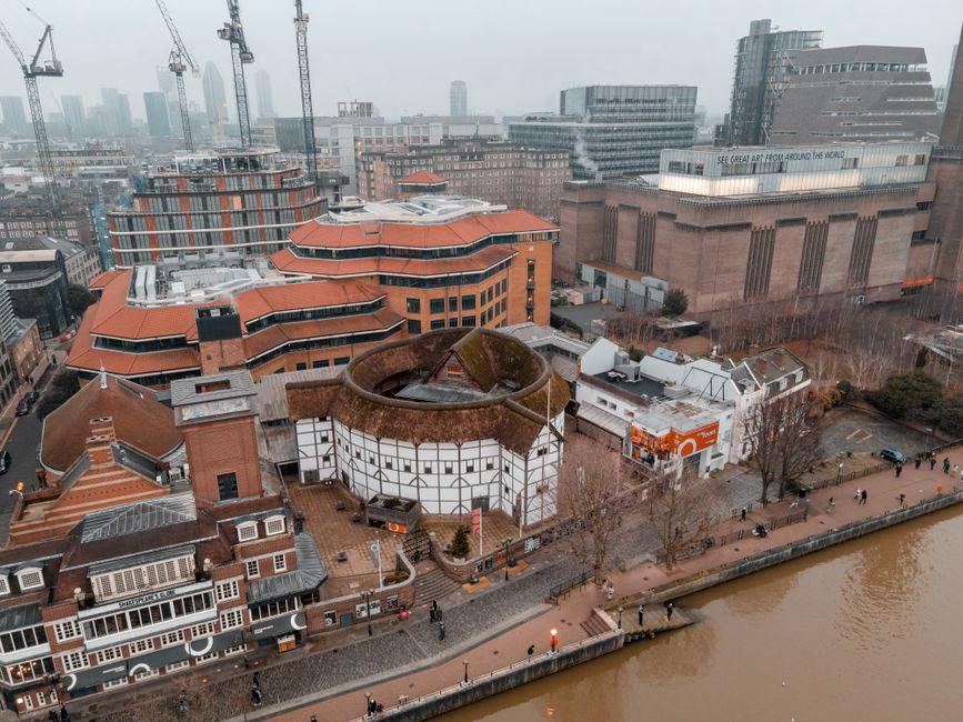 An aerial view of a cityscape featuring a mix of architectural styles, including a distinctive white and black timber-framed building with a circular, moss-covered roof in the foreground, situated along a riverbank with a paved walkway and a few pedestrians. Surrounding structures include modern office buildings with glass facades, some under construction with cranes visible, and older brick buildings with red-tiled roofs. The scene is taken under overcast weather, with diffused lighting highlighting the textures of the buildings, rooftops, and river. In the background, high-rise buildings form a dense urban skyline, with construction cranes indicating ongoing development. The proximity of various building types reflects urban regeneration, which often involves clearing and replacing older structures, aligning subtly with services provided by companies like Rubbish Clearance Hammersmith, who facilitate private waste handling and on-site clearance to support urban redevelopment and maintenance activities.