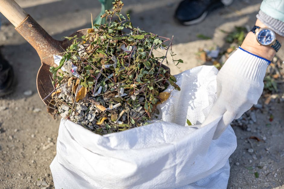 A person wearing a white glove and a wristwatch is lifting a large white waste collection bag filled with garden debris, including a mixture of soil, small rocks, and green leafy plants. The bag appears to be made of woven plastic material with a slightly crumpled texture. The garden waste contains twigs, cuttings, and dry leaves, some of which are partially visible on the top of the heap. A rusty shovel with a wooden handle is partially visible on the left side of the image, resting on the ground which is composed of loose dirt and small gravel. The scene is outdoors, with a focus on the waste collection process, reflecting a typical garden clearance activity that a company like Rubbish Clearance Hammersmith might undertake, emphasizing private on-site clearance or waste disposal services that focus on garden or green waste removal. The lighting suggests natural daylight, highlighting the textures and details of the debris and equipment, creating a clear and professional depiction suitable for accessible viewing and SEO relevance in rubbish removal contexts.