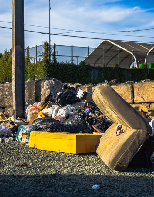A roadside scene featuring a pile of discarded rubbish including black plastic garbage bags, a large yellow plastic container, and an overturned, worn-out beige car seat with visible fabric upholstery and wear marks. The rubbish is spread across a rough asphalt surface, with small scattered debris surrounding the main pile. Behind the waste, there is a low stone wall constructed of irregularly shaped, weathered stones, and a tall metal pole with horizontal wires extending from it, indicating utility infrastructure. In the background, a green hedge partially obscures a tennis court or sports facility with a curved, semi-transparent roof structure, under a partly cloudy sky with patches of blue and scattered clouds. The scene suggests a casual, off-site rubbish collection area, potentially managed by an independent disposal service such as Rubbish Clearance Hammersmith, reflecting a temporary or unmanaged waste accumulation point typical of private waste clearance activities.