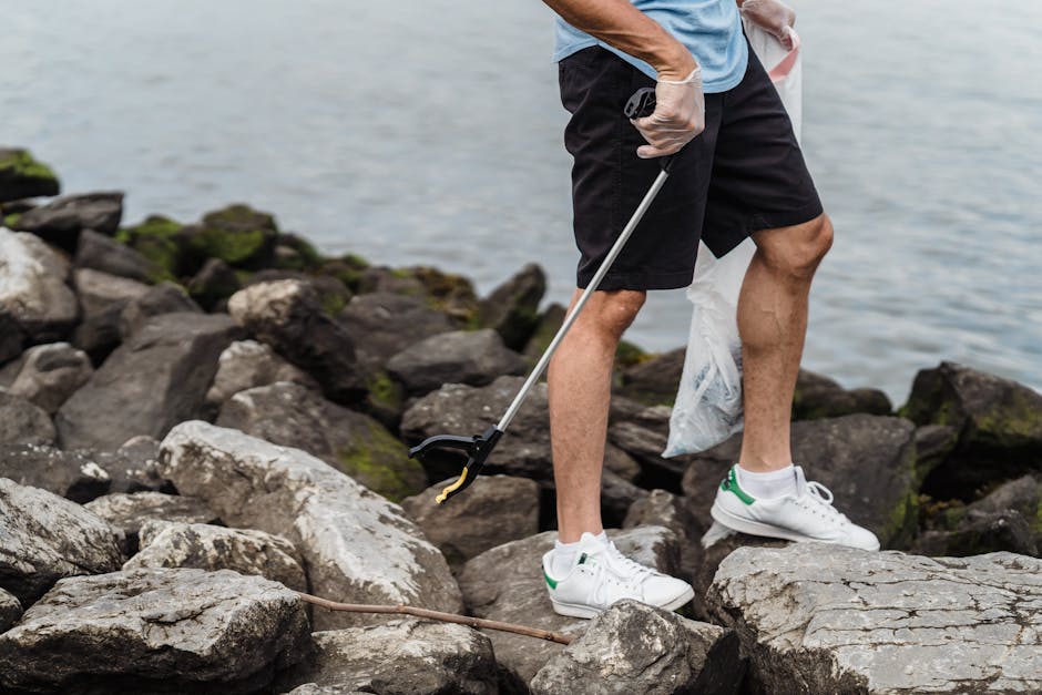 A person wearing black shorts and a light blue t-shirt stands on a rocky shoreline near a body of water, participating in a cleaning activity. They are holding a litter picker in their right hand and wearing white gloves, indicating they are collecting waste from the rocks. Their left hand holds a small plastic bag, likely for gathering trash. The rocks around their feet are large, irregular in shape, and have varying shades of gray and brown, some with patches of green moss or algae. The environment appears natural with moss-covered rocks and calm water in the background, suggesting an outdoor setting near the coast or riverbank. The scene emphasizes private or independent rubbish removal efforts in a scenic outdoor location, as part of environment cleanup activities. Rubbish Clearance Hammersmith offers similar services for rubbish removal from outdoor and urban areas, supporting sustainable waste disposal solutions.